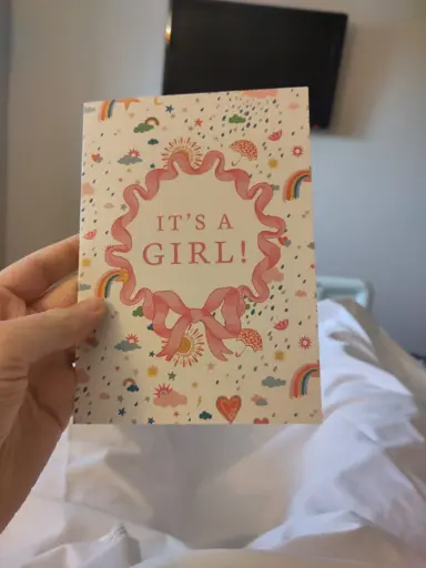 Photo of a card being held by someone in a hospital bed. It reads "It's a girl!" With a pink ribbon, confetti and rainbows surrounding it